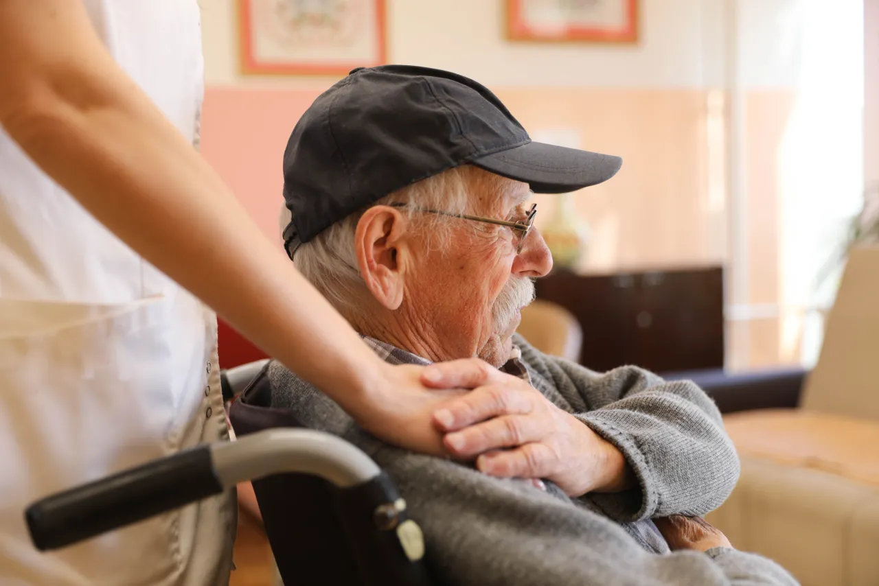 A caregiver's hand resting supportively on an elderly man's shoulder